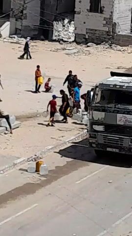 The video shows a group of people, including children, in an outdoor, dusty environment with damaged buildings in the background. Several individuals are carrying or collecting water in large plastic jugs and yellow containers. Some are walking towards or away from a truck that appears to be stopped on the side of a road. The overall scene suggests a situation where people are in need of essential resources like water.