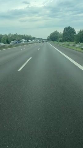 The video shows a highway scene from the perspective of a vehicle driving on it. The sky is overcast with clouds. On the left side of the highway, there are multiple lanes with various vehicles, including trucks and what appear to be campers or RVs. In the distance, there is traffic congestion. On the right side of the highway, there is a shoulder and a guardrail. Trees and greenery line the sides of the highway. A bridge or overpass is visible in the distance. The road surface is asphalt, and lane markings are clearly visible. The overall impression is of a moderately busy highway on a cloudy day.