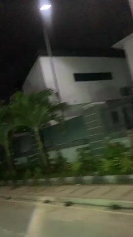 A blurry video captures a street scene at night. The camera is moving, and the footage is shaky. On the right side of the frame, a building with vertical lines and palm trees is visible. On the left, a street with a white line is present. A silver car is seen driving in the distance. The video also shows a digital clock displaying 11:59. Another car, this one black, passes by in the foreground. The scene appears to be in an urban or suburban area with some vegetation and modern architecture. The lighting is dim, consistent with nighttime.