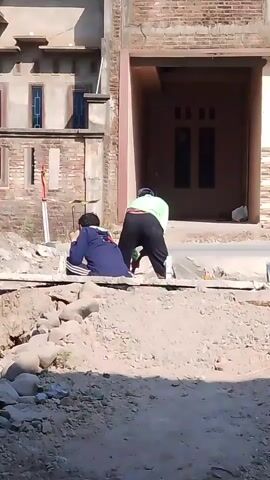 The video shows two individuals in an outdoor construction area. One person, wearing a blue shirt and dark pants, is sitting on a raised platform made of planks. The other person, wearing a green shirt and black pants, is bending over and appears to be working with a tool, possibly mixing or spreading cement. In the background, there is a partially constructed building with brick walls and windows. A motorcycle passes by briefly. The ground is covered in dirt, rocks, and construction debris. The scene is lit by daylight, suggesting it is daytime.