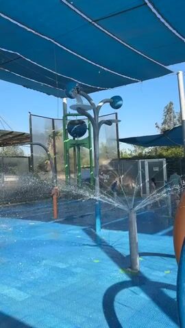 The video shows a water park play area with blue rubberized ground. In the foreground, a gray pole with multiple water sprayers shoots arcs of water into the air. To the right, a large blue and orange tube structure is partially visible. In the background, a blue canopy shades the area. Behind the canopy, a metal fence encloses the play area, and trees and buildings can be seen beyond it. A large green structure with spherical elements is also visible in the background. There are several people in the background, some of whom appear to be children playing in the water. The overall scene depicts a sunny day at an outdoor water play area.
