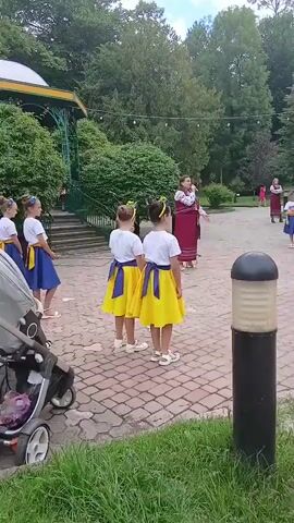 A group of young girls in matching yellow and blue dresses are gathered in an outdoor area, likely a park or a public square. They appear to be performing or rehearsing for a performance, as one girl in the center is singing into a microphone while gesturing. The other girls are standing in formation, some facing the singer, others observing. In the background, a woman in traditional clothing is also present, and another group of girls in similar attire can be seen further away. The environment is lush with trees and greenery, and a gazebo-like structure is visible to the left. A stroller is parked in the foreground, indicating the presence of a caregiver. The ground is paved with cobblestones.
