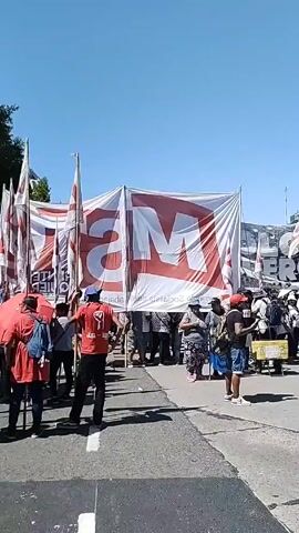 Here's a description of the video:

The video shows a group of people gathered on a street, likely a demonstration or protest. Many are holding flags with a red and white striped pattern. A large banner with the letters "MAS" is visible in the background. People in the crowd are wearing casual clothing and some are carrying backpacks. One person is holding a blue and white beach umbrella. The backdrop features a building with architectural details and green trees. The sky is clear and blue. The video seems to be taken during daylight.