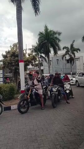 A group of people on motorcycles are lined up on a paved road. The person in the foreground, on a black and yellow motorcycle, is wearing a light-colored jacket and shorts and is looking to the left. Behind them, other people are seated on motorcycles, some wearing helmets and headscarves. A tall palm tree is visible to the left of the frame, and other trees and buildings are in the background. The sky is overcast.