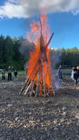 A group of people are standing around a large bonfire that is burning intensely. The fire is made of logs stacked in a teepee shape and is producing a lot of flames and smoke. Some of the people are children and are standing closer to the fire, while others are adults and are standing further away. The scene appears to be outdoors, possibly in a forest or park, with trees and a blue sky visible in the background. The ground is covered in dirt and rocks. One person on the left side of the frame is waving their hands in the air.