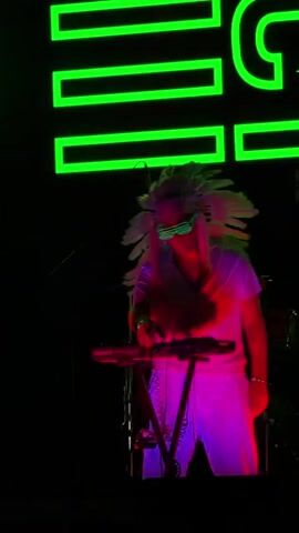 A person wearing a white headdress with feathers, white clothing, and neon green glasses stands behind a keyboard. The person is playing the keyboard and the lighting changes from purple to pink and then to green. Behind the person, a large neon green sign with block letters is visible. The background is dark.