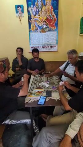 The image depicts a group of six men gathered around a table, seemingly engaged in a game of cards. The setting appears to be an indoor space, possibly a common room or a shop, with a warm yellow-toned wall and a framed picture of a deity on the upper left. A large calendar with an image of Lord Shiva and other figures hangs prominently on the back wall, bearing the text "MODERN BOOK SHOP" at the bottom.

The men are dressed in casual attire, some wearing traditional caps. The table is covered with a patterned cloth, and several decks of cards and what appear to be small bills are spread out. One man in the center is lighting a cigarette with a lighter, while another in the foreground is holding a hand of cards, looking intently at them. A smartphone lies on the table, its screen illuminated. The atmosphere seems relaxed and focused on the game. The lighting suggests it could be daytime, as there's no strong artificial light source visible and the overall illumination is moderate. There are no obvious indicators of weather conditions. The presence of the "MODERN BOOK SHOP" text on the calendar, combined with the location context of Naggar, India, suggests the scene is taking place within or near a bookshop in Naggar. The expressions of the men are generally neutral or contemplative, consistent with concentration during a game.