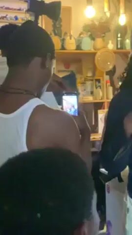 The video shows a crowded indoor space with multiple individuals. The focus is primarily on a few young men. One is wearing a white tank top and another has on a white T-shirt with a cartoon bear print on the back. Someone with long dreadlocks wearing a beanie interacts with them. People are moving around and seem to be socializing in a casual setting. Shelves with various items and hanging light bulbs are visible in the background. At least one person is recording the event with their mobile phone.