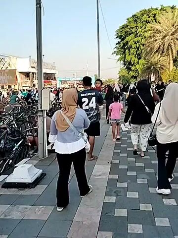 The video opens with a street view, showing a busy sidewalk on the right with several people walking, including a woman carrying a child on her shoulders. On the left, a large number of bicycles are parked, and people are gathered around them. The scene transitions to a wider view of a public square or park, where a large crowd has gathered, mostly consisting of people with bicycles. Some individuals are standing, while others are seated on their bikes. In the background, there are monuments or decorative structures that resemble large, curved tusks or horns, and some flags are visible. A fountain can be seen in the distance. The sun is out, and the sky is clear. The video then shows various people cycling or standing with their bikes, suggesting an event or gathering related to cycling.