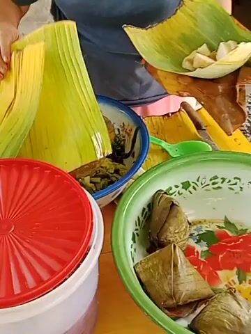 The video shows a close-up of a table setting with food and serving dishes. In the foreground, a large red plastic lid covers a white container. To the right, a green-rimmed bowl contains several parcels wrapped in banana leaves. Behind these, a blue-rimmed metal bowl holds a small glass dish with a spoon in a red sauce, surrounded by green vegetables. A person’s arm is visible on the right side of the frame, reaching towards the blue-rimmed bowl. Banana leaves are scattered around the table, suggesting an outdoor or rustic setting. The lighting is natural, indicating daytime.