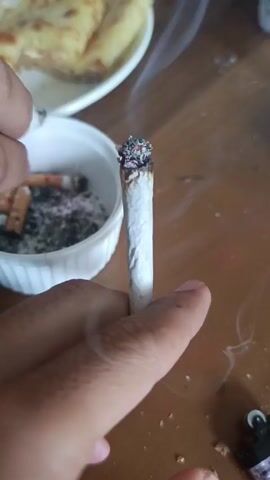 A hand holds a lit rolled cigarette with a white paper wrapping, embers glowing at the tip, and smoke rising. To the left is a small white container with cigarette butts and ashes. In the background, a white plate holds what appears to be food items. The scene is on a wooden surface, with a lighter visible in the lower right corner. The hand holding the cigarette is in focus.