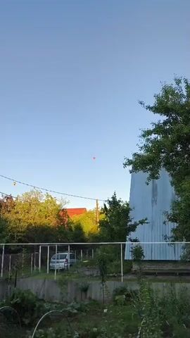 The video shows a clear blue sky with a single, small, orange hot air balloon ascending. Below the balloon, the tops of trees and rooftops of houses are visible, along with power lines stretching across the frame. The scene appears to be an outdoor residential area.