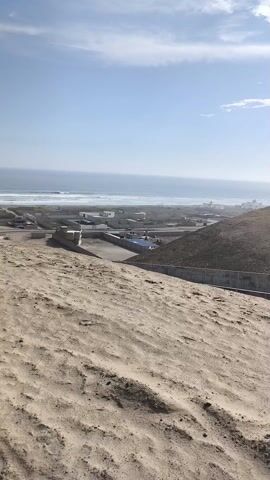 The video shows a view from a sand dune overlooking a coastal area. In the distance, the ocean meets the horizon under a bright, slightly hazy sky. White waves crash on the sandy beach. In the middle ground, there's a collection of white buildings and what appear to be concrete structures or ponds. The foreground is dominated by the sandy dune, textured with patterns of wind and erosion. There's a concrete wall partially visible on the right side of the frame, likely part of the structure below. Overall, the scene is a combination of desert landscape meeting the coast with visible signs of human construction. The wind can be heard in the audio.
