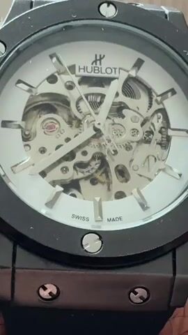 The video shows a close-up, high-angle, eye-level, stationary shot of a black and white skeleton watch. The watch has a black casing and a white face with silver hour markers and hands. The watch's internal mechanisms are visible through the transparent face. The brand name "HUBLOT" is visible at the top of the watch face, with the "H" logo above it. Below the watch hands, "SWISS MADE" is printed on the face. The watch is resting on a dark surface, and the bottom edge of a person's hand holding the watch is visible at the very bottom of the frame.