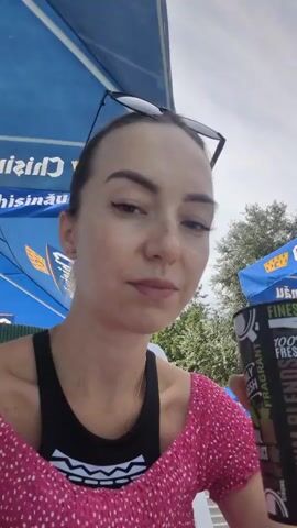 A woman with blonde hair, wearing sunglasses pushed up on her head, a pink polka dot top over a black tank top, and light blue nail polish, is drinking from a black can with white and green text. Her eyes are closed. She is outdoors, with a blue umbrella overhead and foliage in the background.