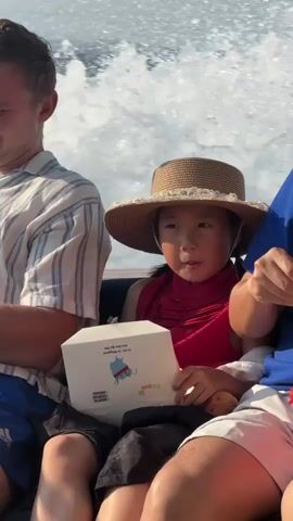A young girl wearing a straw hat and a red top sits on a boat, holding a white box. She looks down at the box, her mouth slightly open. To her left, a person wearing a striped shirt sits with their arm resting on their lap. To her right, another person wearing a blue shirt is visible, holding a white utensil. The boat is moving, creating a wake in the blue water behind them. The sky is visible in the background, and there appears to be land in the distance.