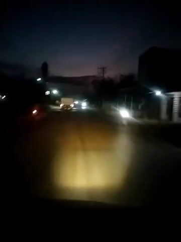 The video shows a view from inside a vehicle driving on a road at night. The headlights of the car illuminate the road ahead, revealing a dark asphalt surface. In the distance, the taillights of a truck are visible. Streetlights line the sides of the road, casting a soft glow. A tall structure, possibly a minaret, is seen against the dark sky, suggesting the location may be a town or village with a mosque. The sky has a hint of twilight. Utility poles are visible along the roadside.