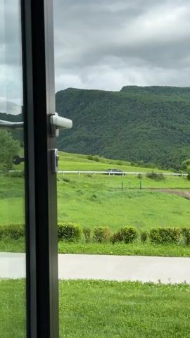 From a slightly ajar door, a scenic view unfolds. A meticulously manicured lawn, defined by short, uniform green grass, meets a concrete path just beyond the door frame. Further into the distance, a sprawling green field stretches towards rolling hills covered in lush, verdant trees. A white fence, partially obscured by the tall grass, runs horizontally across the field. A dark-colored vehicle is parked on a dirt path adjacent to the fence. The sky above is overcast with grey clouds. The door frame is black, and the door handle and lock are silver. A key hangs from the door.
