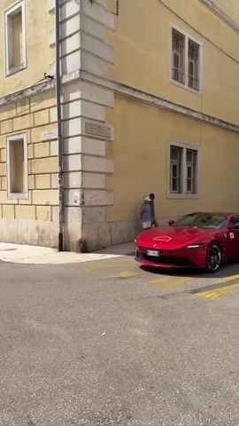 A red Ferrari drives down a cobblestone street lined with aged, yellow buildings. Two people stand near the corner of a building as the car passes. The car has a white circular sticker with the number 113 on its side. Another red car is visible further down the street, as is a white van. People are walking on the sidewalk in the distance. The sky is partly cloudy.
