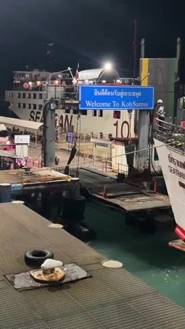 This media file depicts a nighttime scene at a ferry terminal in Nathon, Thailand. The primary subjects are several large ferries docked at the pier, with their lights illuminated against the dark sky. One ferry, prominently featured on the right, has the name "SEATRÄNFERRY 6" visible in large lettering. A blue sign overhead reads "ยินดีต้อนรับสู่เกาะสมุย" in Thai, followed by "Welcome To Koh Samui" in English, indicating the destination.

The setting is a busy port at night, with a visible loading ramp connecting the shore to the ferries. Several people are present, including a ferry worker in a high-visibility vest standing on the deck of the "SEATRÄNFERRY 6" and other figures visible on the ferries and the dock. The water is visible between the ferries and the pier.

The activity appears to be the arrival or departure of ferries, with passengers or vehicles likely preparing to board or disembark. The atmosphere is functional and operational, illuminated by artificial lights. The presence of multiple large vessels and the welcoming sign suggest a significant transportation hub. No specific weather conditions are apparent beyond it being dark. The visible text clearly identifies the location as Koh Samui and the ferry service.