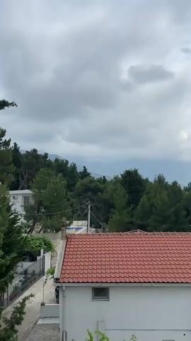 The video presents a panoramic view of a coastal landscape under a cloudy sky. It begins looking straight ahead over dense green trees and a red-tiled roof towards distant, hazy mountains. The camera then slowly pans to the right, gradually revealing more of the scene.

As the pan continues, several white residential buildings with terracotta roofs become visible in the foreground, nestled among abundant greenery including tall, slender cypress-like trees and broader pine-like trees. White outdoor stairs descend on the right side of the frame.

The midground opens up to show a vibrant coastline with a light-colored beach stretching along a tranquil bay. Beyond the beach, a city extends along the coast and inland, characterized by numerous buildings and urban structures. The calm, blue-green sea is dotted with what appear to be small boats or vessels. Majestic mountains form the backdrop to the city and bay, receding into the cloudy distance. The overall weather appears overcast or partly cloudy during daytime.

The scene depicts an expansive view of Šušanj, Montenegro, showcasing its residential areas, lush vegetation, coastal features, and the surrounding mountainous terrain.