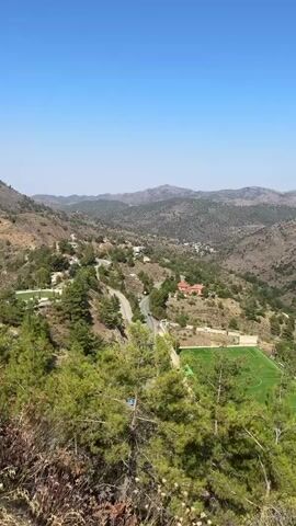 The video shows a panoramic view of a mountainous landscape under a clear blue sky. In the foreground, there are pine trees and some dry foliage. In the mid-ground, a winding road cuts through the terrain, with a few buildings scattered along it. A soccer field with green turf and white markings is visible on the right side of the image. The background consists of rolling hills and mountains covered with sparse vegetation. The overall scene depicts a rural, possibly Mediterranean, environment. No people or specific actions are discernible in the video.
