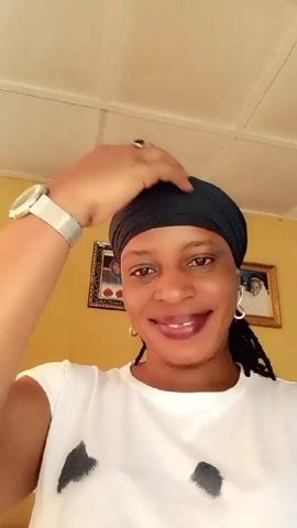 A woman wearing a black headwrap and a white shirt poses for a selfie video. She is wearing a gold-colored watch and silver infinity-shaped earrings. Her hair is in braids. She smiles and adjusts her headwrap with her right hand. Two framed pictures are on the mustard-yellow wall behind her. The ceiling is white and has a grid pattern.
