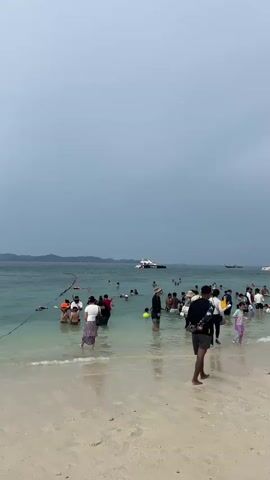 The video shows a crowded beach scene with many people wading in the shallow ocean water. The water is clear and a turquoise color near the shore, becoming deeper blue further out. In the background, several boats are visible, including a white yacht and smaller speedboats. The sky is overcast and a light gray color. People are dressed in casual beach attire, with some wearing swimwear and others in t-shirts and shorts. Some individuals are holding bags or towels. A few people are standing on the sandy shore, while the majority are in the water, interacting with each other or simply enjoying the ocean. The overall atmosphere appears to be one of a busy tourist destination on a cloudy day.