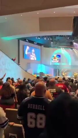The video shows a church service with a preacher on stage. A band is playing behind him, and the audience is watching and listening. There are two large screens on either side of the stage showing the preacher. The church is brightly lit with colored lights. People in the audience are wearing casual clothes and sports jerseys. Smoke fills the room at one point.