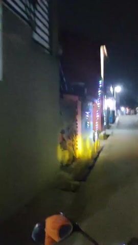 The video displays a low-light, blurry perspective from the viewpoint of someone riding a motorcycle at night. The scene appears to be a street or alleyway lined with buildings. Decorative lights, predominantly pink, green, and blue, are strung along the sides of the buildings, suggesting a festive or celebratory atmosphere. The buildings themselves are simple structures, some with visible windows and doorways. The road surface is visible, and the motorcycle's headlight illuminates a portion of it ahead. There are no clear views of people or specific actions, as the motion and low light create a generalized impression of movement through a decorated, nocturnal environment.