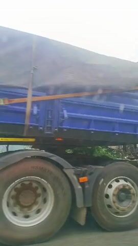 The video shows a large white truck driving down a road, with a blue truck partially visible next to it. The white truck has the text "MEGA MARKET (PVI) LTD." and "NO UNAUTHORISED PASSENGERS" on its side. The camera then pans to show trees and a grassy hill next to the road, with a cloudy sky above.