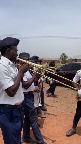 In a daytime outdoor scene, a band consisting primarily of men and some women is playing brass instruments on a dirt road. The musicians are dressed in matching uniforms: white shirts, dark blue trousers or skirts, and dark caps. Many of them are playing trombones and trumpets. The individuals appear focused on their instruments as they move or stand. In the background, simple buildings and sparse trees are visible under a clear, bright sky, suggesting a rural or semi-urban setting. Parked cars are also seen further back, with a woman in a dark dress walking past one. The overall atmosphere indicates a public event or procession, likely in Ladura Foron, Nigeria, given the context. An emblem is visible on the shirt of the band member closest to the camera, though specific text or details are not discernible.
