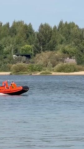 An orange and black inflatable boat with two people aboard is moving across a body of water. The boat is creating a wake behind it. In the background, there is a shoreline with trees, vegetation, and some buildings. The sky is clear and blue.