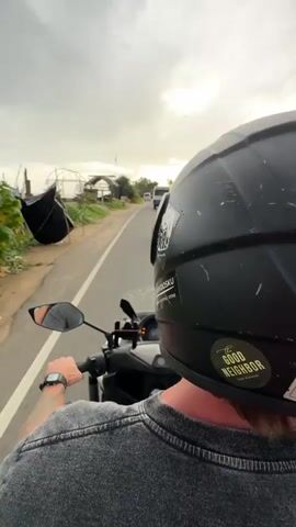 The video shows a first-person perspective of someone riding a black motorcycle on a paved road. The rider is wearing a black helmet with stickers on the back and a grey shirt. The rider's left hand is on the handlebars, and a black smartwatch is visible on their wrist. In front of the motorcycle, there are other vehicles on the road, including a white van and several scooters. To the left of the road, there is a grassy area with some structures and vegetation. The sky is cloudy with sunlight breaking through. The overall environment appears to be a coastal road.