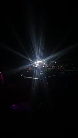 A nighttime outdoor concert is depicted. Bright stage lights illuminate a band performing on a raised stage. A crowd of people is visible in front of the stage, silhouetted against the lights. Cars are parked in the foreground, also in silhouette. The overall atmosphere is dark with dramatic lighting.