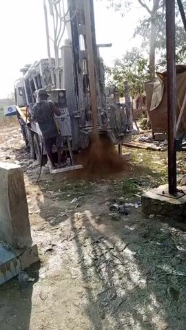 A man operates a drilling rig on a truck in a muddy, sunny outdoor area. The rig is actively drilling, ejecting a cloud of brown soil and water. Debris litters the ground, and there is a temporary shelter nearby. The man, wearing a tank top, stands on the truck's rear platform operating the machinery.  Trees are visible in the background. A concrete barrier is on the left side of the frame.