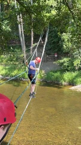 A man wearing a blue t-shirt, black shorts, and a red helmet is walking across a narrow rope bridge suspended over a shallow, clear river. He is attached to a safety harness and is holding onto ropes for support. Another person, only their red helmet and shoulder visible, is in the foreground looking towards the man. The bridge is made of two parallel ropes with wooden slats, and is supported by additional ropes and cables attached to trees on either side of the river. The environment appears to be a wooded area with lush green vegetation and trees. The water is shallow enough to see the rocky riverbed below. The sun is shining, creating dappled light on the water.