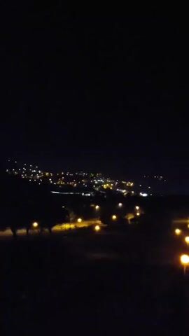 The video shows a night view of a town from a slightly elevated perspective. The sky is dark with the moon visible. Below, numerous lights of the town are scattered across the horizon, with rows of streetlights running along roads in the foreground. Silhouetted trees frame the scene.