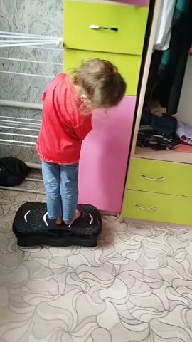 The video shows a young child standing on a black balance board. The child is wearing a red t-shirt and blue jeans. The child is facing a tall piece of furniture that appears to be a dresser or wardrobe. The furniture has green and pink drawers. The floor is covered with a patterned linoleum. In the background, there is a drying rack with white bars. The child moves around on the balance board and looks at the camera. The video zooms in on the child's face at the end.