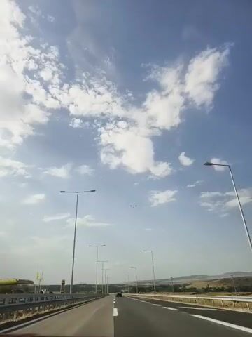 The video shows a first-person perspective of a car driving on a highway. The sky is blue with scattered white clouds. There are streetlights lining the highway, and guardrails on the side. In the distance, there are hills. A few birds are visible in the sky.