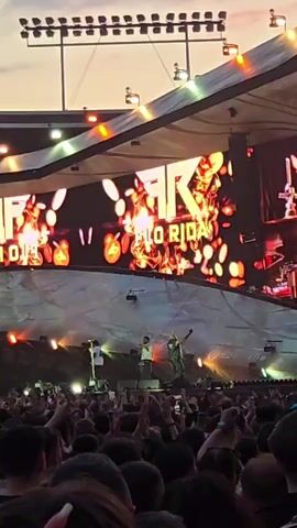 A wide shot shows a large outdoor concert crowd facing a stage. A massive LED screen above the stage displays the words "FLORIDA" and the letters "FKR". The screen is illuminated with abstract, fiery graphics. On the stage, two performers are visible in silhouette. The crowd is densely packed, with many people holding up their phones, likely to record the event. The sky above is a soft twilight color, and stage lights are visible.