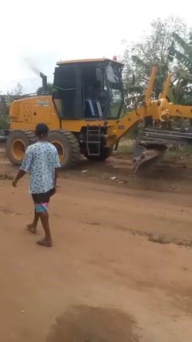 A man in shorts and a printed shirt stands on a dirt road in front of a large, yellow XCMG road grader. The grader appears to be moving forward, smoothing the surface of the road with a large blade positioned beneath it. The sky is overcast, and trees are visible in the background.