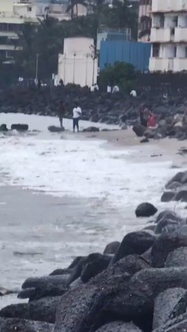 The video shows a rocky shoreline with waves breaking on the beach. Several people are visible on the beach and near the water's edge. Buildings and palm trees are in the background. The overall atmosphere appears to be a public recreational area by the sea.