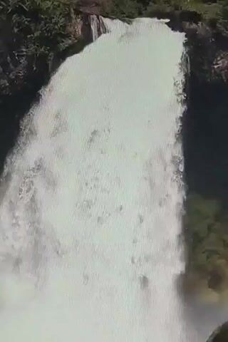 This video shows a close-up, aerial view of a large waterfall. The water is white and frothy as it cascades down a dark rock face covered in lush green vegetation. The camera is angled slightly down, providing a sense of the immense power and scale of the waterfall. There are no people or man-made objects visible in the frame. The lighting suggests it is daytime, with sunlight illuminating the water. The sound of the rushing water is a prominent feature of the video.