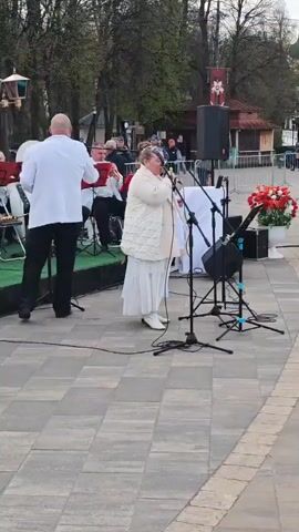 The video captures a scene from an outdoor event, possibly a local celebration, set on a paved square.

In the foreground, a woman dressed in white is singing into a microphone. To her left, musicians are playing instruments, including an accordion. 

Across from the stage, a small group of people are gathered near a tent set up with DJ equipment. 

In the background, people are walking around, some wrapped in Russian flags. The environment includes trees, buildings, and a hint of a river in the distance.

The general atmosphere suggests a festive gathering, with live music and people enjoying the public space.