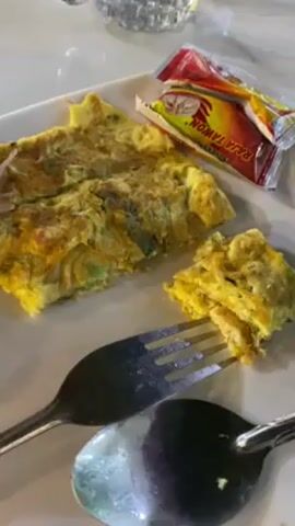 The video shows a white square plate with a partially eaten omelet, a small packet of Raja Tawon brand sauce, and a fork and spoon on a white marble surface.  A tall glass of a green drink is also on the table.  The omelet appears to contain vegetables. The fork is holding a piece of the omelet. A partially filled glass of water is also visible in the background.