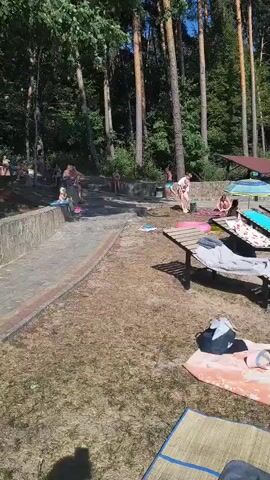 The video shows a sunny day at a lake or beach. There are many people present, some sunbathing on loungers and towels, while others are swimming or walking near the water. A wooden pier extends into the lake, with people on it and in the water around it. There are trees in the background, suggesting a natural, wooded area. There is also a small covered structure, possibly a snack bar or changing area, visible in the distance. The overall atmosphere appears to be relaxed and recreational, with a focus on outdoor activities.
