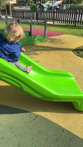A young child with blonde hair, wearing a blue jacket and striped pants, is sitting at the top of a bright green plastic slide in a playground. The child pushes off with their hands and begins to slide down. The playground surface is yellow and there is a partial view of a fence and other playground equipment in the background. The sun is shining.