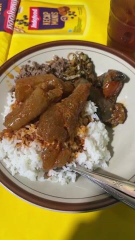 A close-up, top-down shot shows a plate of food on a yellow surface. The plate is brown with a cream-colored rim. It contains a mound of white rice topped with several pieces of meat in a reddish-brown sauce, some minced meat, fried anchovies, and what appears to be pieces of eggplant. A fork and spoon are resting on the right side of the plate. In the background, out of focus, is a yellow bottle with red and white labeling, and a glass of amber liquid.
