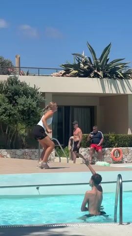 The video shows a sunny day outside near a swimming pool. In the foreground, a young person is in the pool, with their back to the camera. In the mid-ground, a woman in athletic wear is performing an exercise, possibly a jump or a stretch, on the pool deck. She is facing away from the camera and looking upwards. Another person, a man, is sitting on the edge of the pool deck behind the woman, looking towards her. There are also trees and part of a building visible in the background.